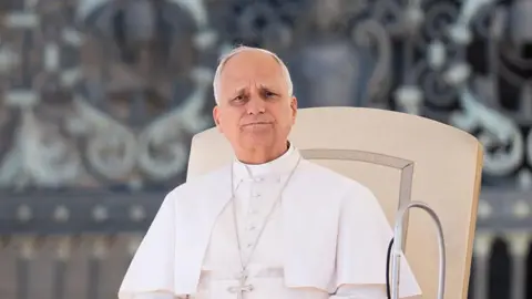Getty Images Pope Leo XIV delivers his speech to the faithful during the Wednesday General Audience in St. Peter's Square at the Vatican on November 5, 2025. He is wearing his traditional white robe and is sitting on a cream-coloured chair. Behind him is a blurred background of an ornate facade at St Peter's Square.