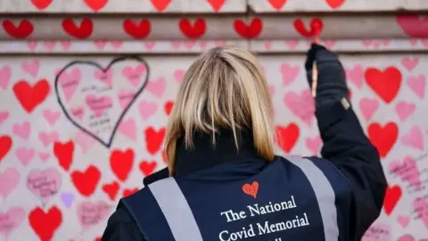 Getty Images A woman paints a heart on a wall as part of the Covid memorial. The grey wall is covered with pink and red hearts. The woman has blonde shoulder-length hair and is wearing a dark coat with a dark blue vest over the top which says "The National Covid Memorial" on it.