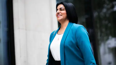 PA Media Shabana Mahmood smiles as she walks past a beige brick building wearing a blue suit and white shirt as she arrives at the BBC building in London on Sunday.