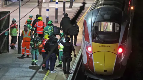 PA Media Emergency workers are stood on a train platform on the left next to a train which is at a standstill. 