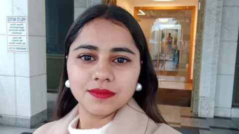 Family handout Harshita Brella looks into the camera for a selfie while standing outside a building. Her hair is open, and she is wearing bright red lipstick and pearl drop earrings.