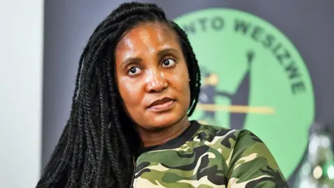 Gallo Images/Getty Images Duduzile Zuma-Sambudla with long braids and wearing a camouflage bomber jacket over a black T-shirt is seen during a MK party media conference in Johannesburg in 2024. Behind her can be see the green and black MK party logo.  