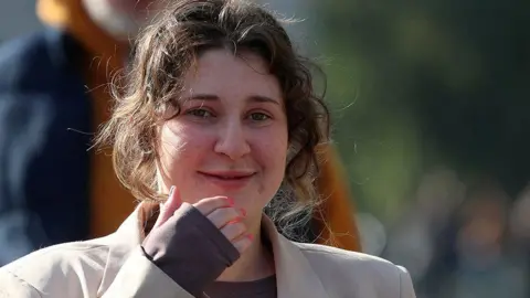 Reuters Bella Culley is walking on the street and smiling. She has short, brown curly hair and is wearing a beige coat and dark top.