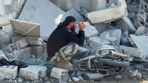 Reuters A man is seen squatting in profile and holding both of his hands together on piles of rubble from a destroyed building. He is bearded and wearing a black top and headgear, camouflage trousers and brown boots; a motorcycle is toppled on its side. 