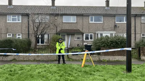 BBC A police officer outside the address in Moredon. It is a terraced property that faces a grassy area. Police tape can be seen across the street.