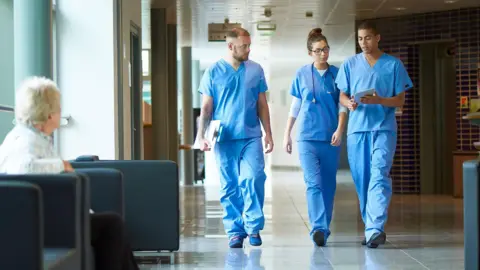 Getty Images Three medics in blue scrubs, two men and a woman, walk along a hospital corridor. One of the men is holding a clipboard, the other some papers and the woman has a stethoscope around her neck. They are walking side by side, engaged in conversation. In the foreground, there is woman sitting on a black cushioned chair. She is turned away from the camera, looking towards the medics.