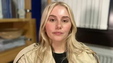 BBC Jayla looking at the camera. She has long blonde hair, brown eyes and a silver hoop nose piercing. She is wearing a cream jacket.
