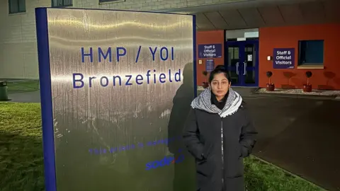 Zarah Sultana MP/X A woman in a long coat and black and white check scarf, standing outside a prison with her hands in her pockets