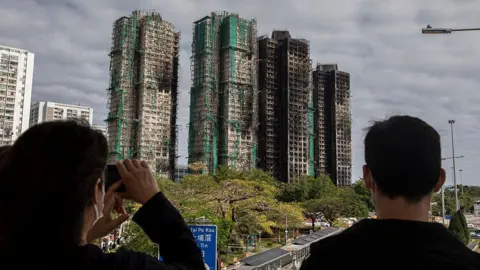 Getty Images The back of two people's heads as they look at four charred high-rise blocks in the distance, with trees and roads between them and the towers