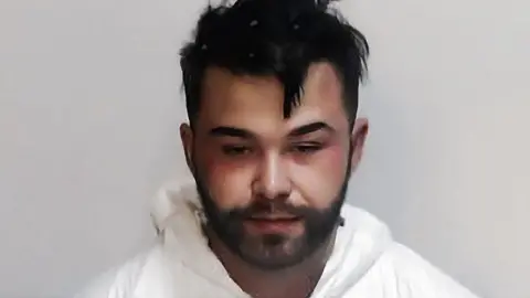 Police Scotland Mugshot of a man in a white forensic-style protective suit with a hood, standing against a plain light gray background. The person has short, dark, slightly messy hair with small white specks visible in the hair