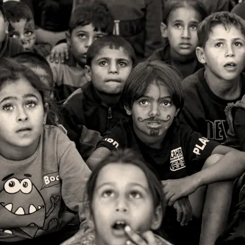 BBC An image in black and white of young children sitting on the ground and looking up, one of them has a beard and moustache drawn on his face in pen