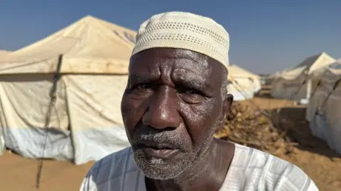 Ed Habershon / BBC Abdulqadir Abdullah Ali, wearing a white cap, looks at the camera, with tents in the background