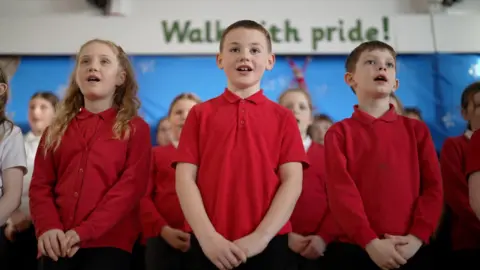 Dan Nelson / BBC Students at Mersey Drive Community Primary School sing in their school hall. It's a close-up shot of three singing primary age children, two boys centre and right, and one girl on the left, all in red school uniform tops. More children stand behind them in rows in the choir. 