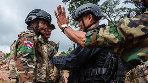 Getty Images Two troops in Cambodian military uniform and helmets face off with a troop in black uniform and helmet, while in the foreground another soldier in military uniform holds out his hand with the palm acing outwards