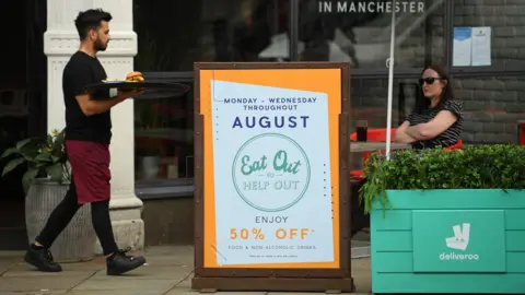 Getty Images A waiter carries a tray with a burger on it to a lone diner who sits under and umbrella in the shade. She has sunglasses on and her arms folded