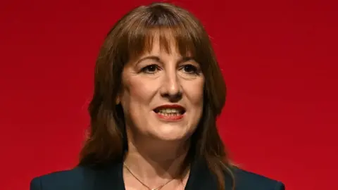 Getty Images Rachel Reeves, who has shoulder length brown hair, in front of a red background. She is wearing a black suit jacket and a gold necklace. 