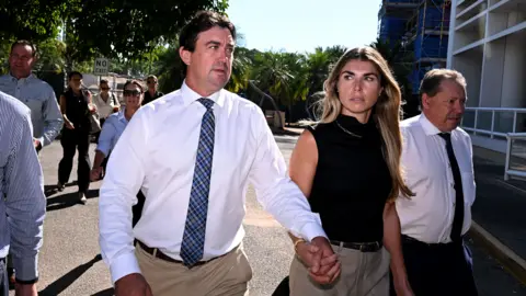 Getty Images A man in a blue check tie and a white shirt arrives at court holding the hand of a blond woman wearing a black top
