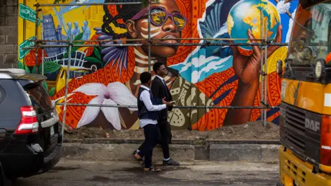 Toyin Adedokun / AFP via Getty Images Two men walk past a brightly coloured mural featuring a woman in sunglasses holding a globe. There is a can and a bus on either side