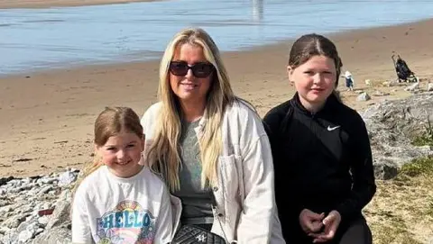 Family photo A young white girl wearing a t-shirt saying hello, her mother and her older sister sit on a rock at the edge of the beach. The mother, Lucy, has long blonde hair and wears black trousers, a plain tshirt and a white light jacket and black handbag over it. She is wearing sunglasses. Lola to her right is aged about 11, is wearing black trousers and sport top. all three are smiling at the camera. It's a sunny day. A few people sit on the beach behind them.