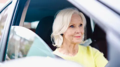 Getty Images An older woman driving a car. She has blonde hair and is wearing a light yellow jumper.