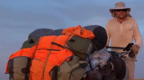 Karl Bushby A man wearing a khaki sunhat and light coloured clothes pushes an overloaded cart containing his possessions and water.