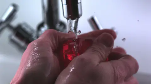 BBC A pair of hands are underneath a tap of running water and are holding a bar of red soap. They are about to lather up the soap.
