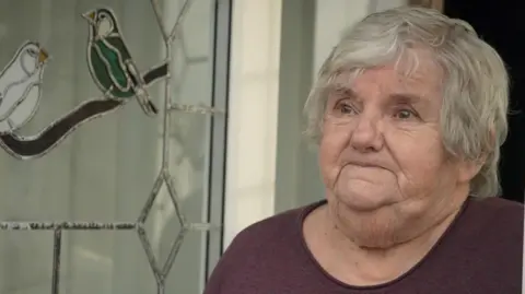 Qays Najm/BBC A woman with short grey hair and a purple top stands beside an open external white uPVC door with a stained glass effect of two birds in the centre.