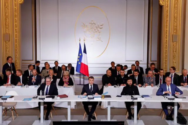 French President Emmanuel Macron, British Prime Minister Keir Starmer, Ukrainian President Volodymyr Zelenskiy, and German Chancellor Friedrich Merz sit at a long white table in front of rows of officials at meeting