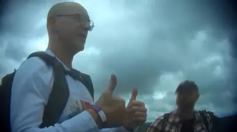 BBC Mr Lawler, a bald man wearing a white long-sleeve t-shirt with a partially-visible graphic on the front, gives a double thumbs-up gesture towards somebody not in frame. He is wearing a rucksack and standing next to another man, whose face is blurred.