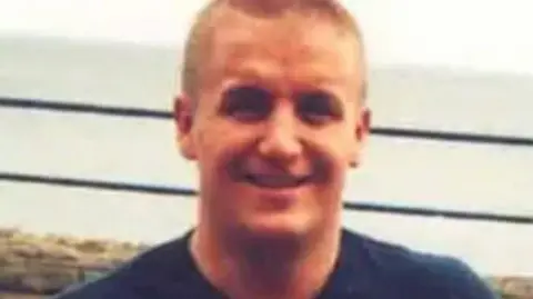 Merseyside Police A picture of a smiling Joseph Cummins, with short blond hair and wearing a blue T-shirt, with a view of the sea visible in the background.