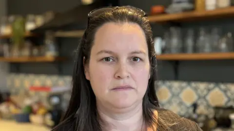 BBC A woman stands in a kitchen. She has long brown hair and is wearing a brown top