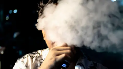 Getty Images An Asian man puffs on a vape, his face mostly obscured by a cloud of vapour