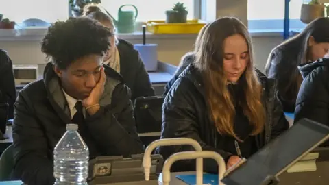 Ann Gannon / BBC In a classroom, two students sit in their coats while working. One male student with black hair is leaning on his hand while in a puffer jacket. He is sat next to a female student who has long mousey brown hair who is writing with a pen on paper and is wearing a parker style coat. 