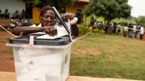 Getty Images A ballot box outside is  in the foreground. A woman in a facemask is placing her vote inside.
