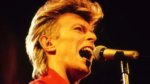 Getty Images David Bowie sings into a microphone on stage. He his bathed in yellow light and is wearing a red top.