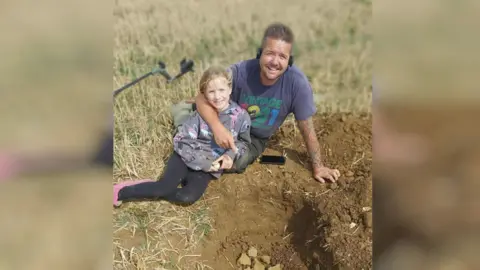Simon Weller A man and his daughter lie on a field. In front of them is a dug hole that has some lumps of metal inside. 